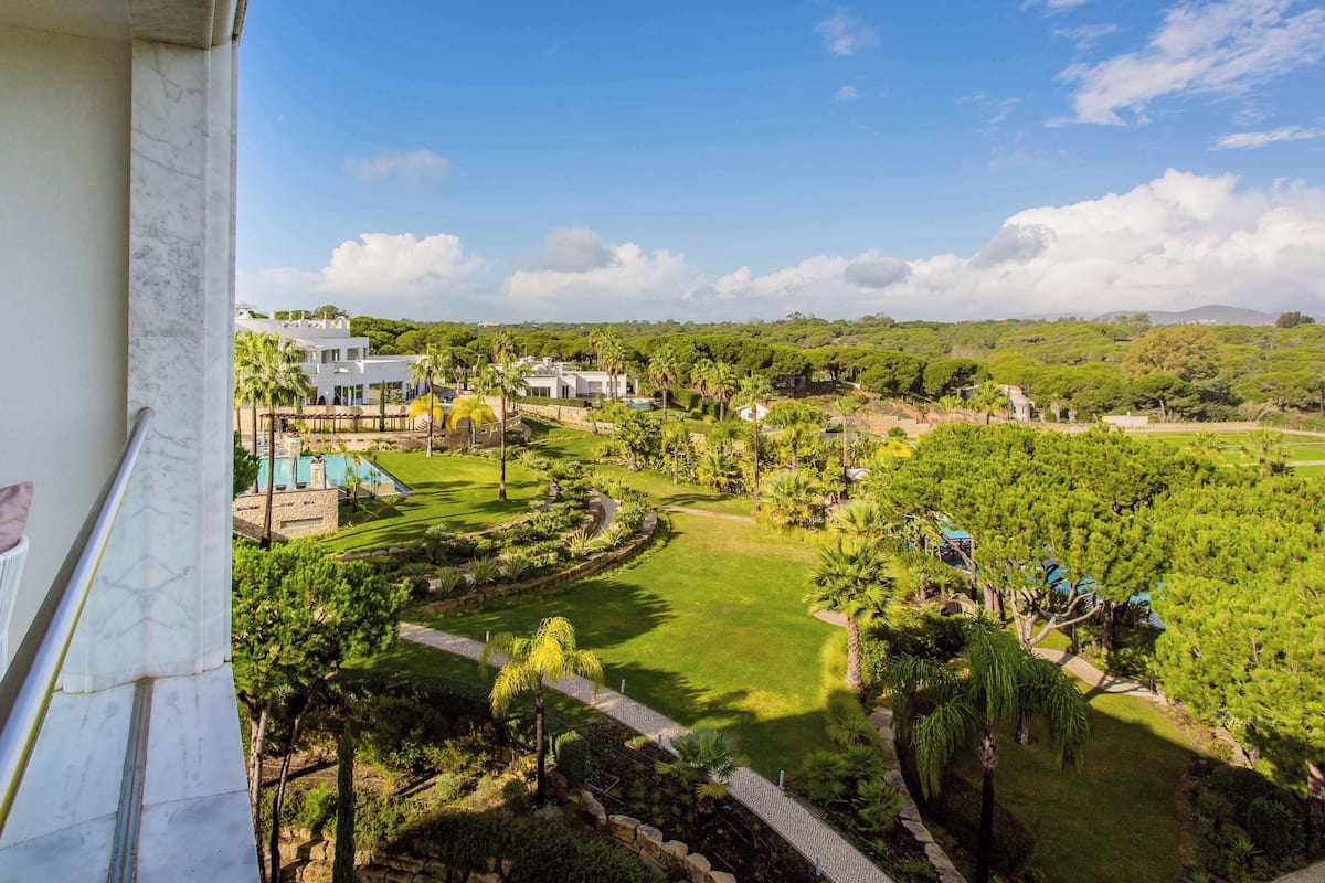 A balcony with a view of a lush garden, a pool, and modern buildings.