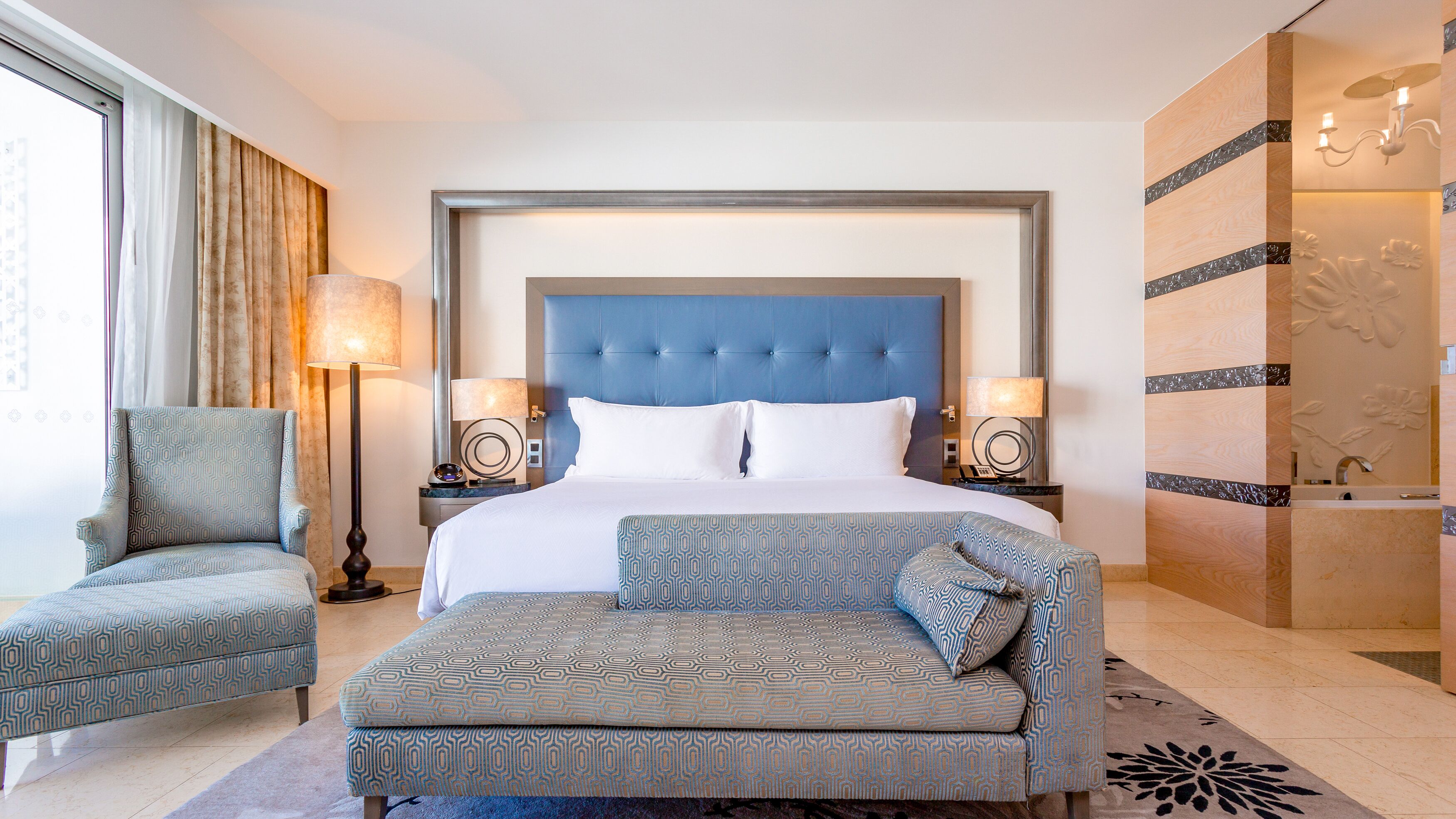 A hotel room with a large bed, two armchairs, a sofa, a nightstand with a lamp, and a wall with a floral design.