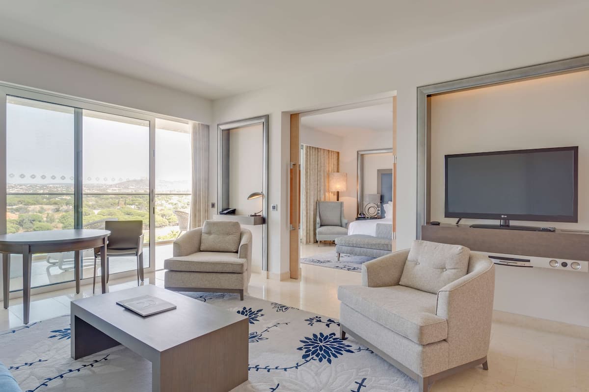 A modern hotel room with a balcony, a flat-screen TV, a dining area, and a living room with a sofa and armchair.