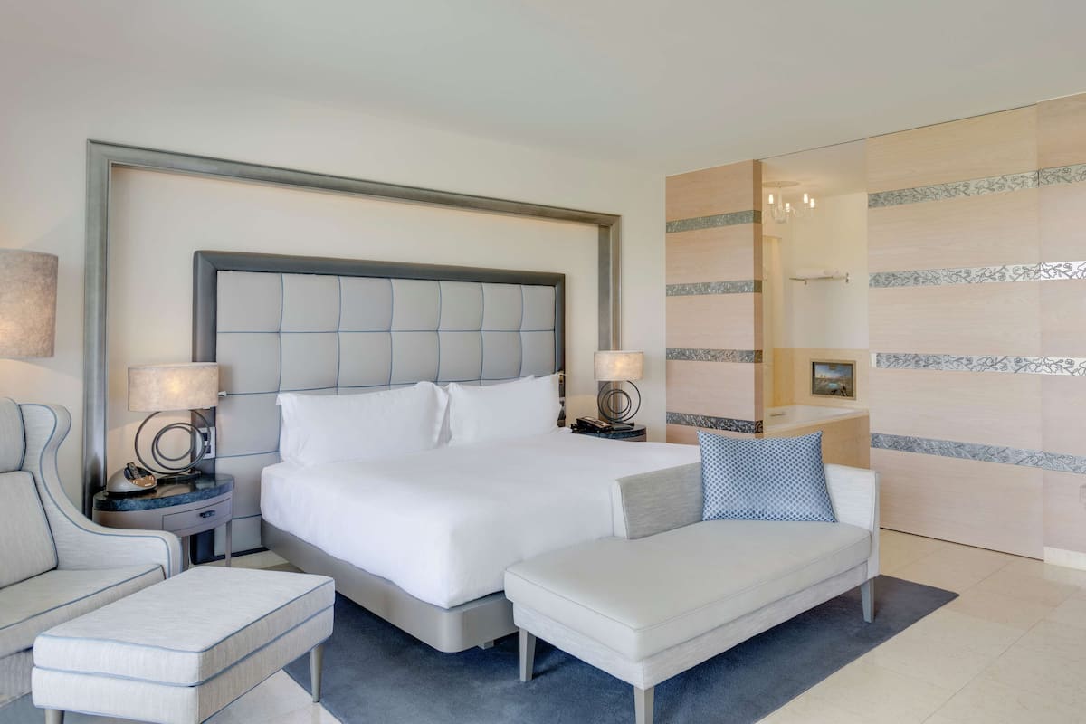 A modern hotel room with a large bed, a bedside table with a lamp, a small sofa, and a bench.