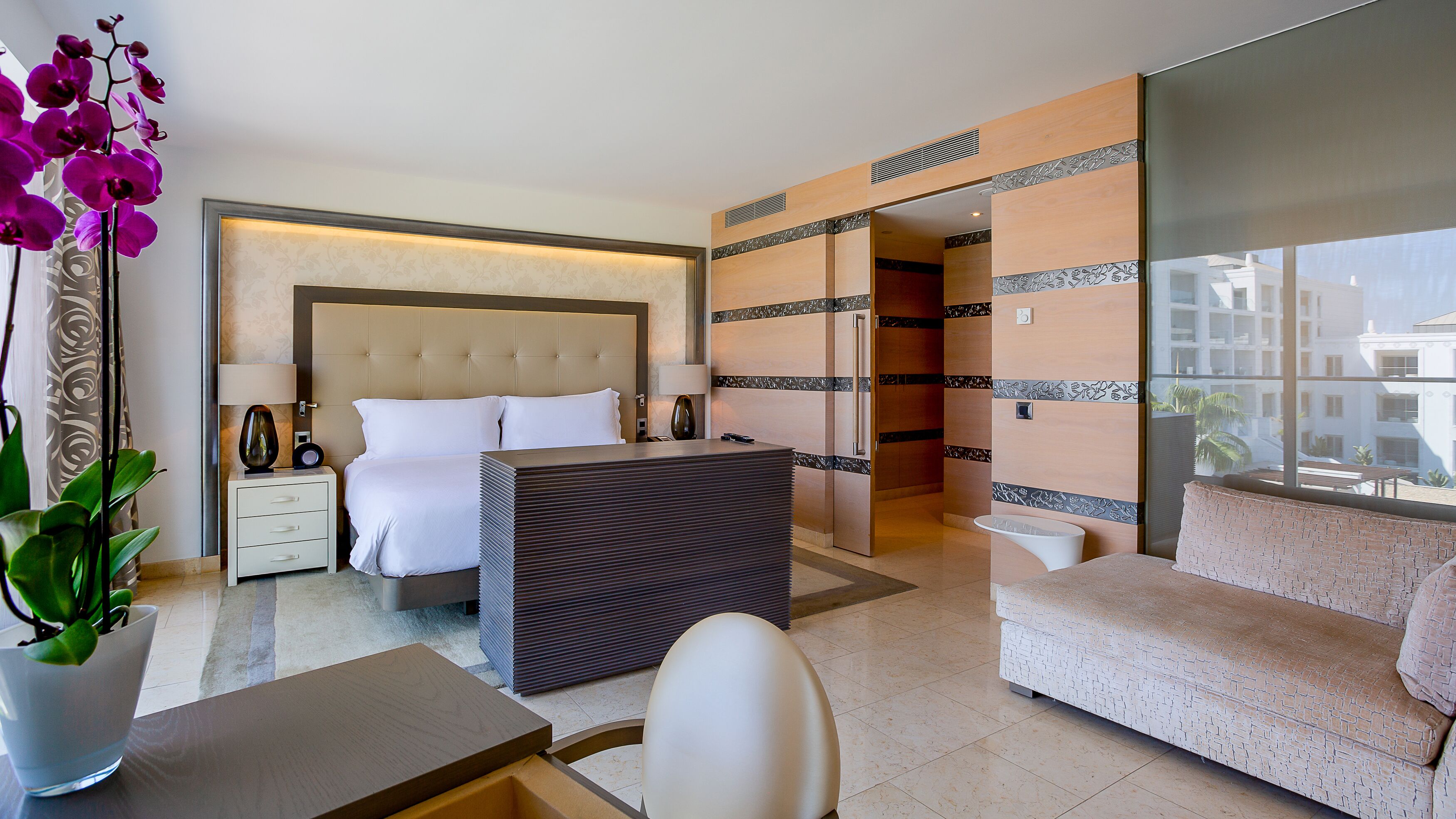 Habitación de hotel moderna con cama, sofá, mesa de comedor, pequeña cocina y un amplio ventanal con vistas a la ciudad.