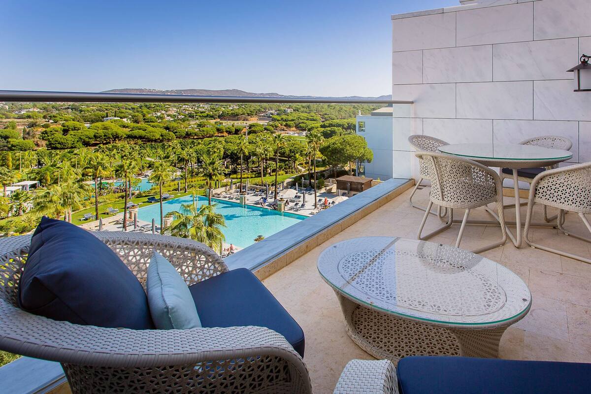 A balcony with wicker furniture, a glass table, and a view of a pool and greenery.