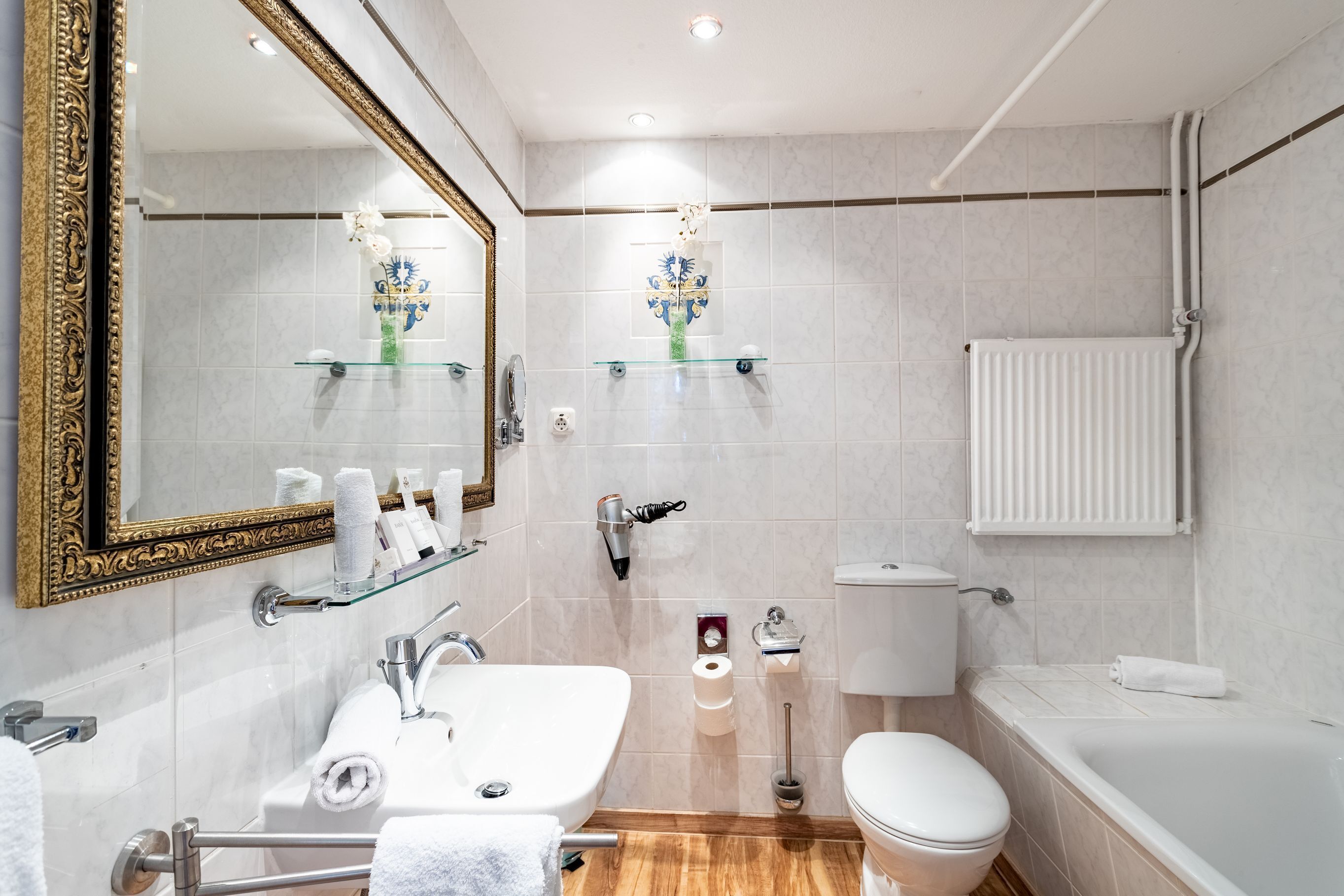 A modern bathroom with a large mirror, white fixtures, a glass shelf with toiletries, a toilet, and a bathtub.