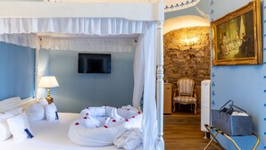 A four-poster bed with white bedding and rose petals, a television mounted on the wall, a stone wall, a painting, and a chair. - Hotel Burg Trendelburg (Trendelburg)