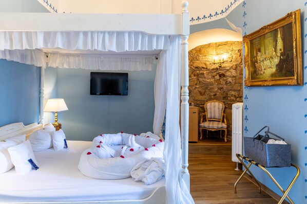 Una cama con dosel, ropa de cama blanca y pétalos de rosa, un televisor montado en la pared, una pared de piedra, un cuadro y una silla.