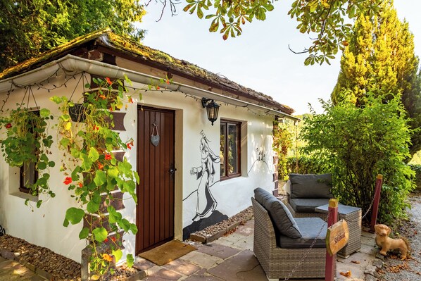 Uma casa pequena com fachada branca, porta marrom e um mural de uma mulher. Um sofá de vime e um cão pequeno estão do lado de fora.