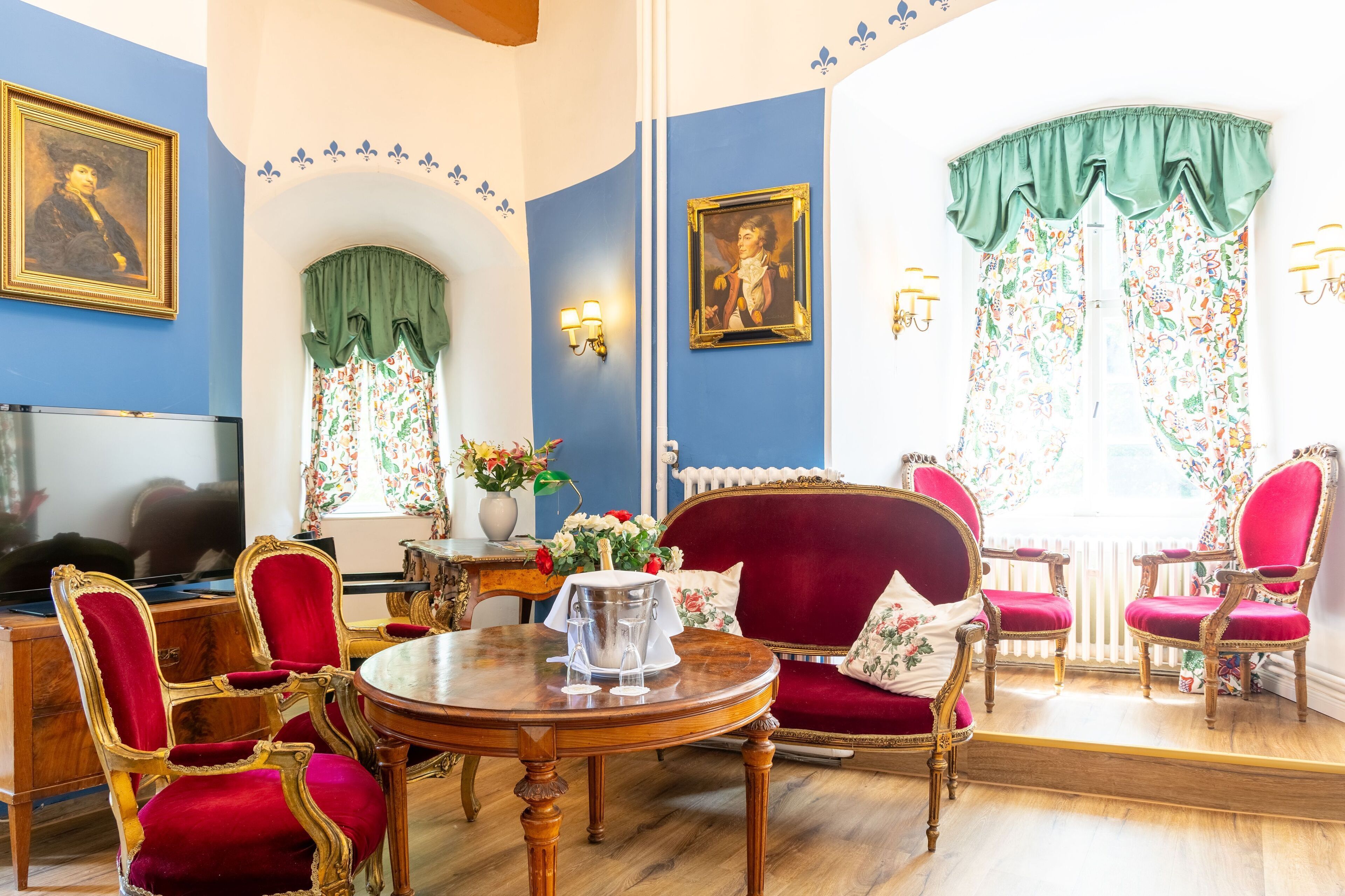 A room with a dining table, red upholstered chairs, a red sofa, and two wall-mounted paintings.