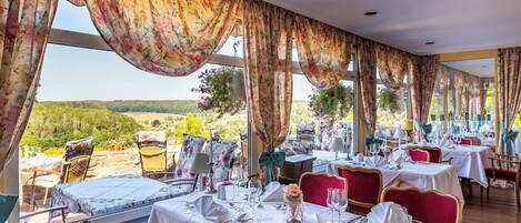 A restaurant with tables set for dining, large windows with curtains, and a view of greenery outside.