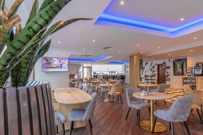 A modern restaurant with round tables and patterned chairs, a TV screen, and a checkered floor.