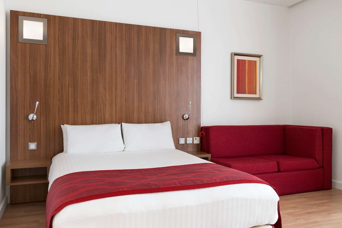 A hotel room with a wooden headboard, a bed with white linens, a red sofa, and a framed picture on the wall.