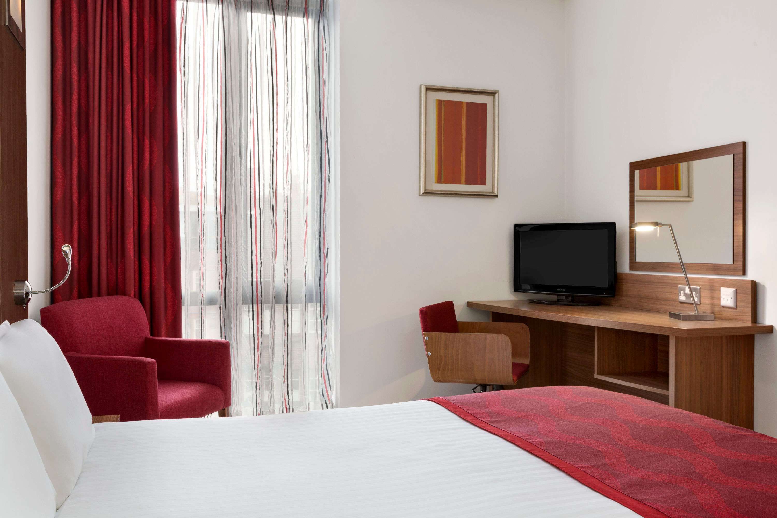 Une chambre d’hôtel comprenant un lit, un bureau avec une télévision, une chaise, une lampe de bureau et un grand miroir.