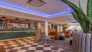Un vestíbulo de hotel con suelo de cuadros, una zona de bar con diversas botellas de licores y arreglos de asientos que incluyen un sofá y sillones.