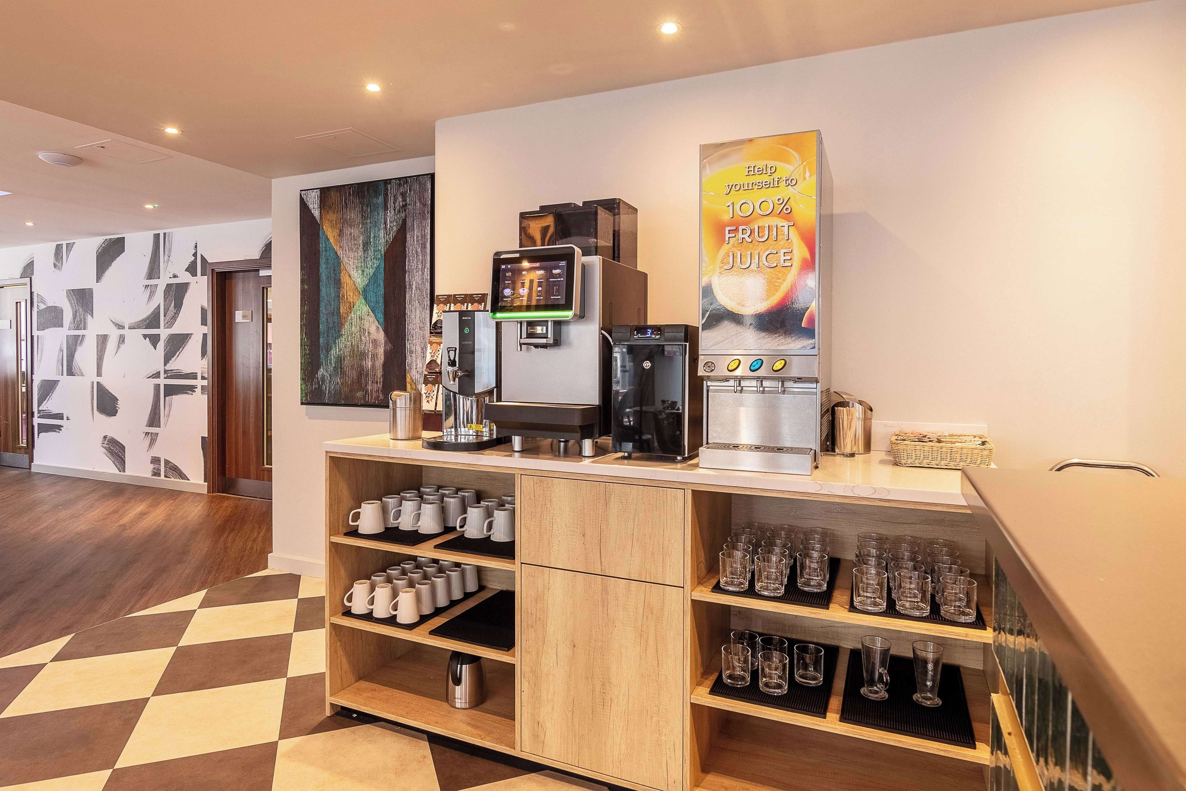 A modern coffee station with a fruit juice dispenser, coffee machines, and a variety of mugs and glasses.