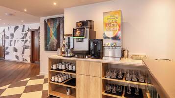 Un espace café moderne équipé d’un distributeur de jus de fruits, de machines à café et d’une variété de tasses et de verres.