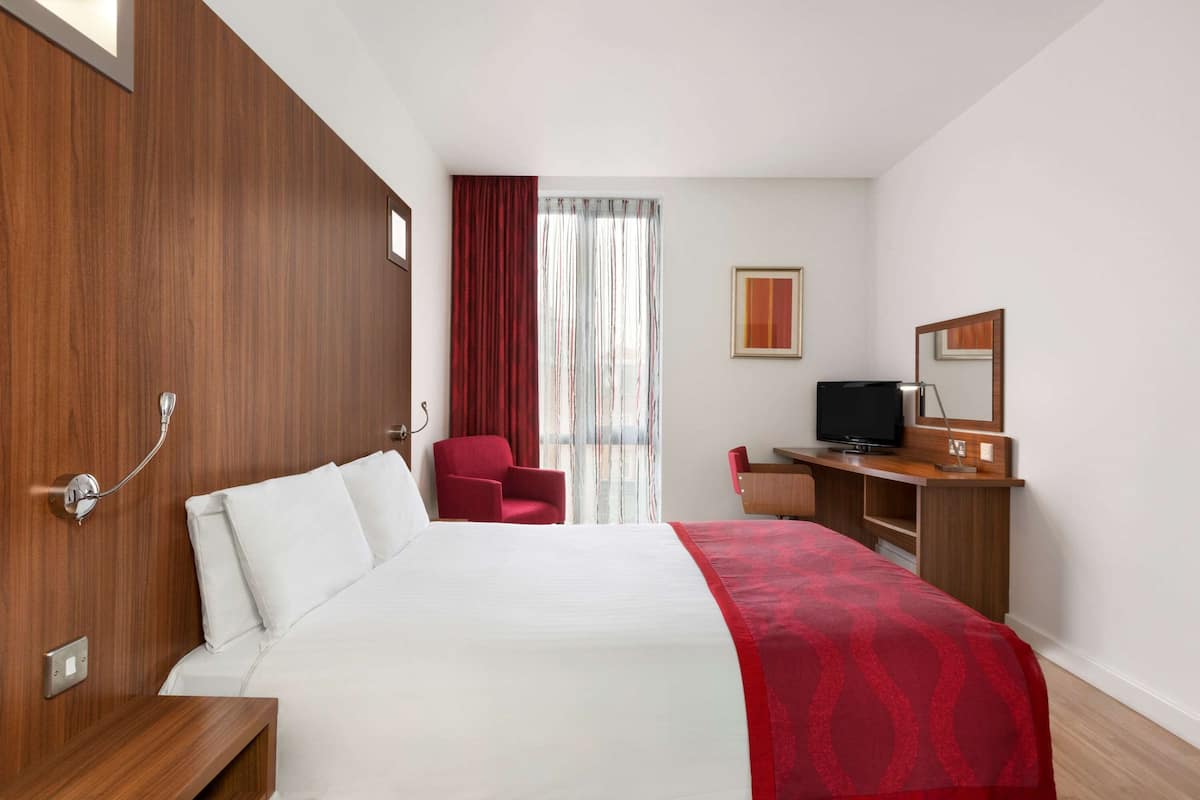 A hotel room with a wooden headboard, a bed with white linens, a red chair, a desk with a TV, and a large window with curtains.