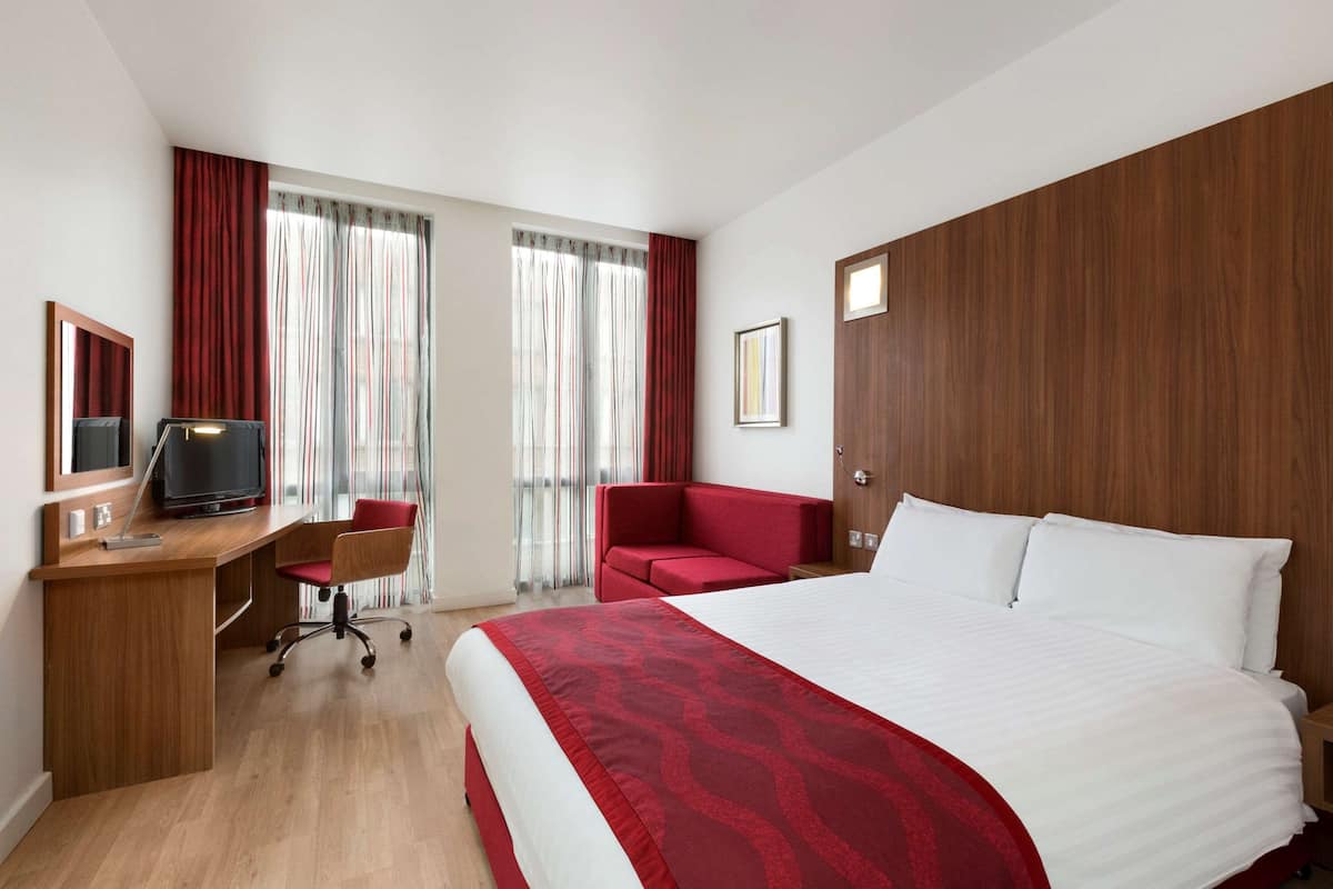 A hotel room with a wooden desk, a red chair, a bed with white linens, and a large window with curtains.