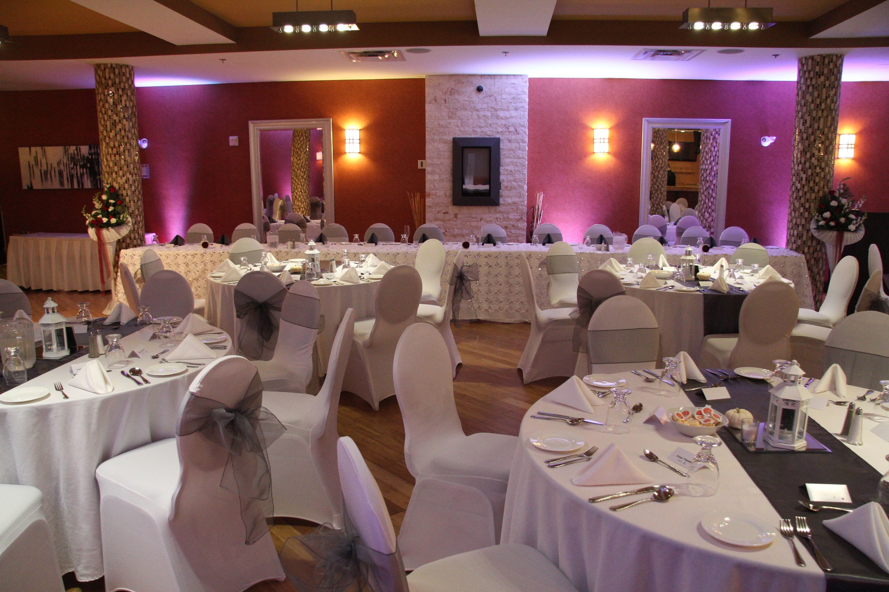 Een balzaal met ronde tafels gedekt voor een formeel evenement, met witte tafelkleden, stoelen versierd met linten en een stenen accentmuur.