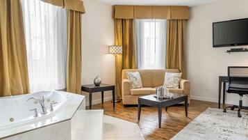 A modern bathroom with a large bathtub, a vanity, and a mirror. There is a beige sofa and a small table with a lamp in the living area. A flat-screen TV is mounted on the wall.