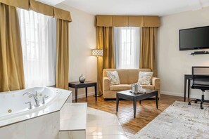 A modern bathroom with a large bathtub, a vanity, and a mirror. There is a beige sofa and a small table with a lamp in the living area. A flat-screen TV is mounted on the wall. - Comfort Hotel Bayer's Lake (Halifax)