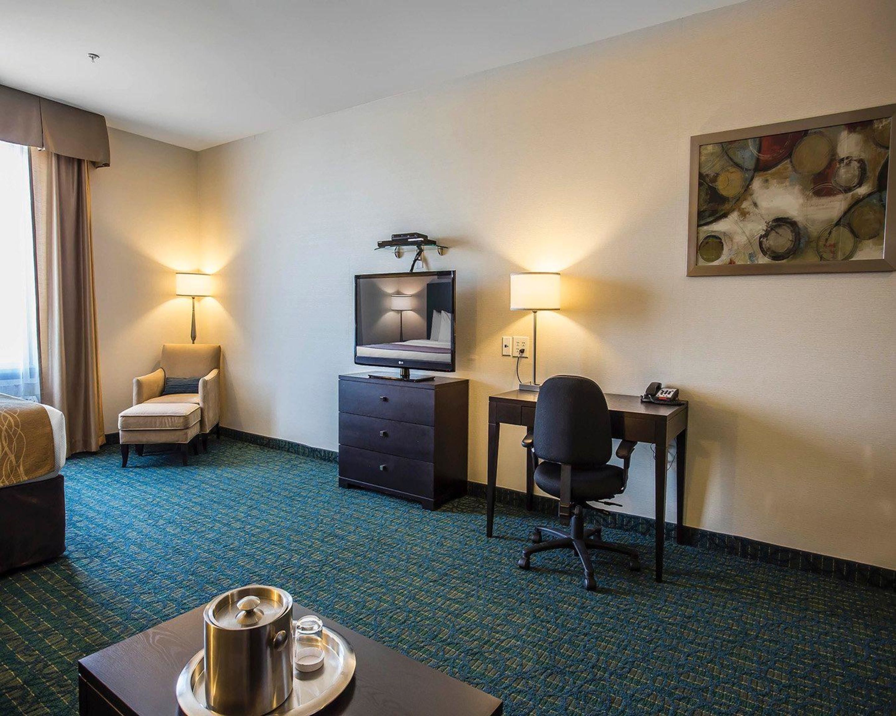 A hotel room with a bed, a desk, a chair, a TV, a lamp, a painting, and a tray with a pot and glasses.