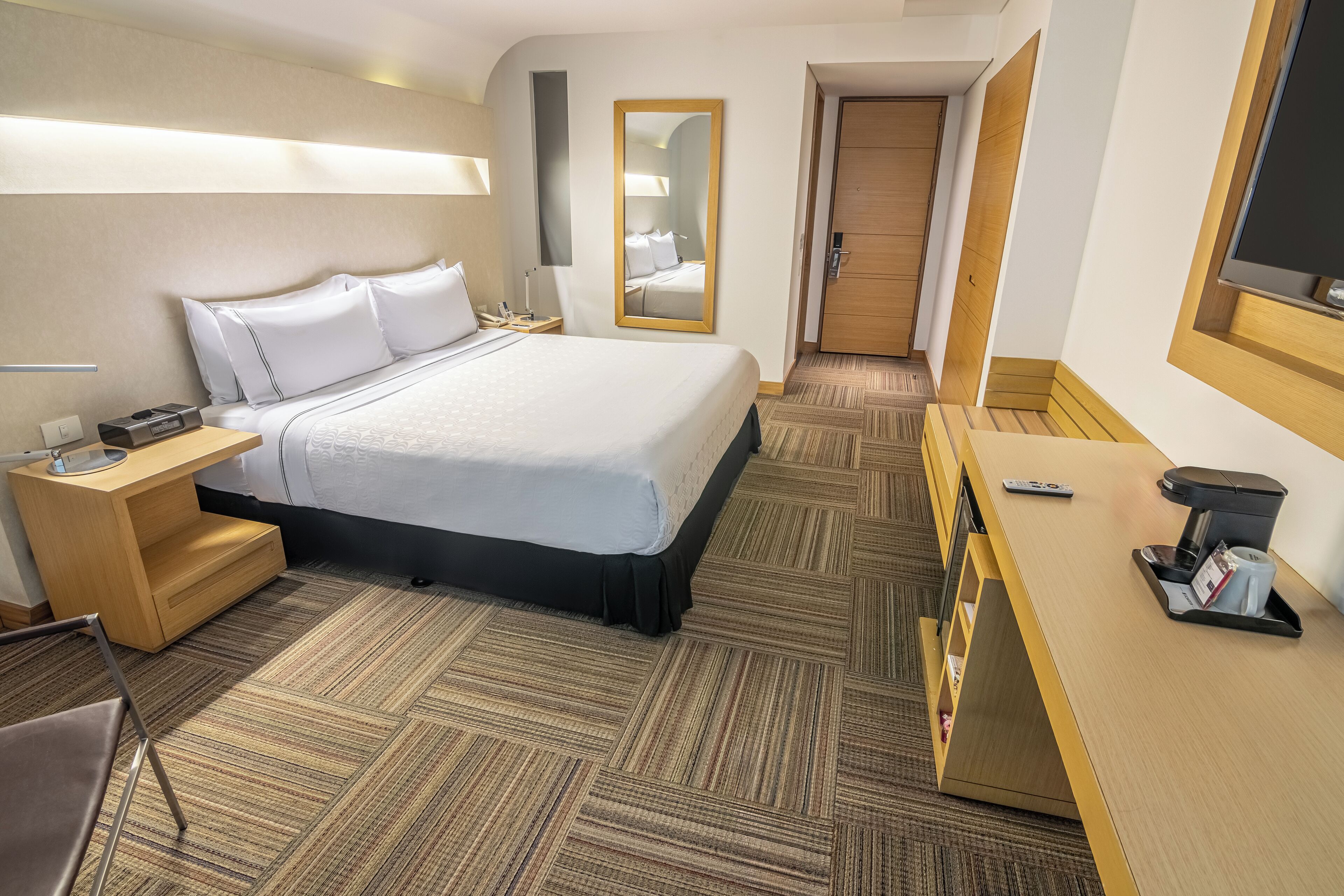 A hotel room with a bed, bedside table, desk, and a mirror.