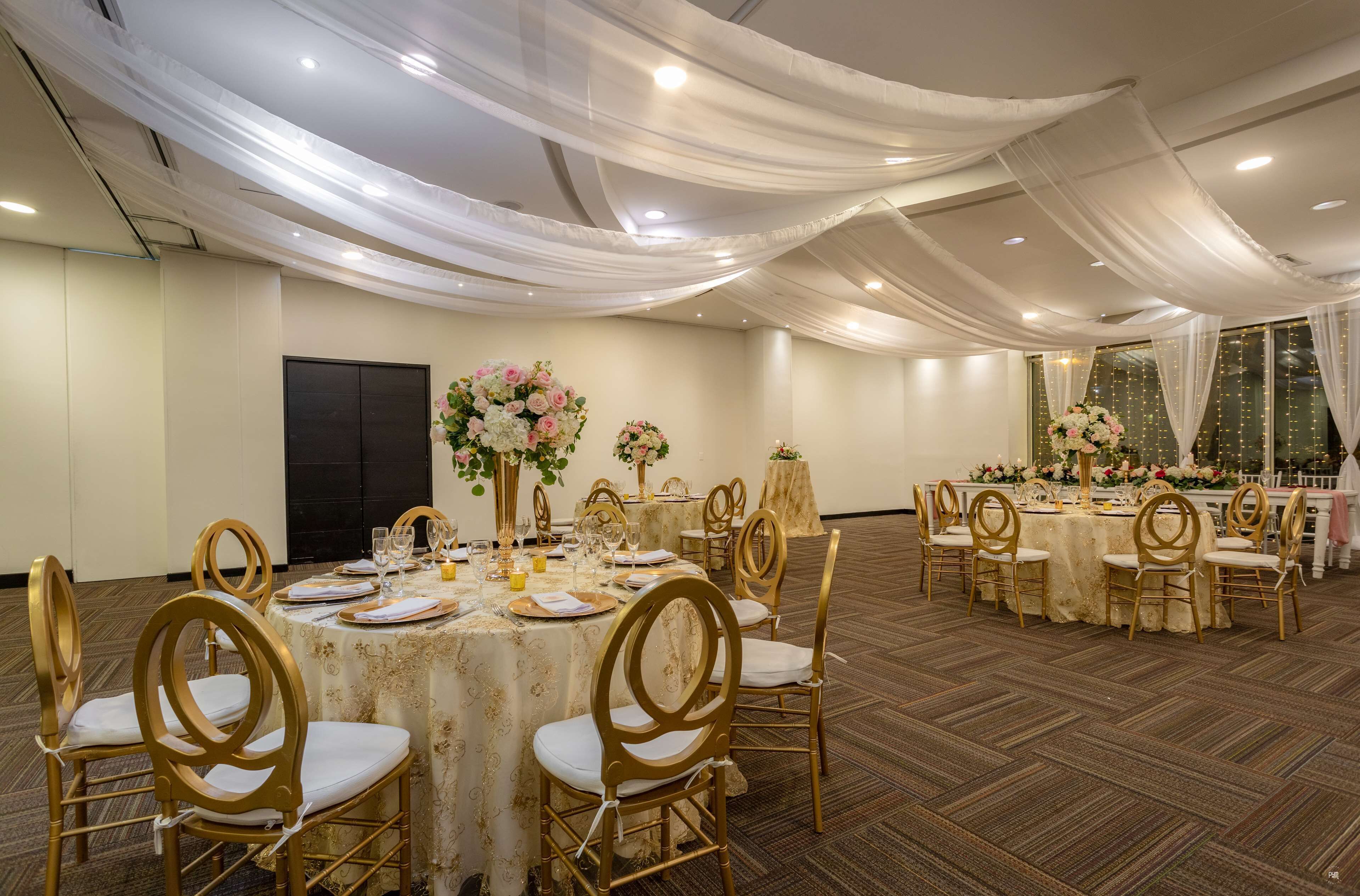 Un salón de banquetes con mesas redondas, sillas doradas y centros de mesa florales.