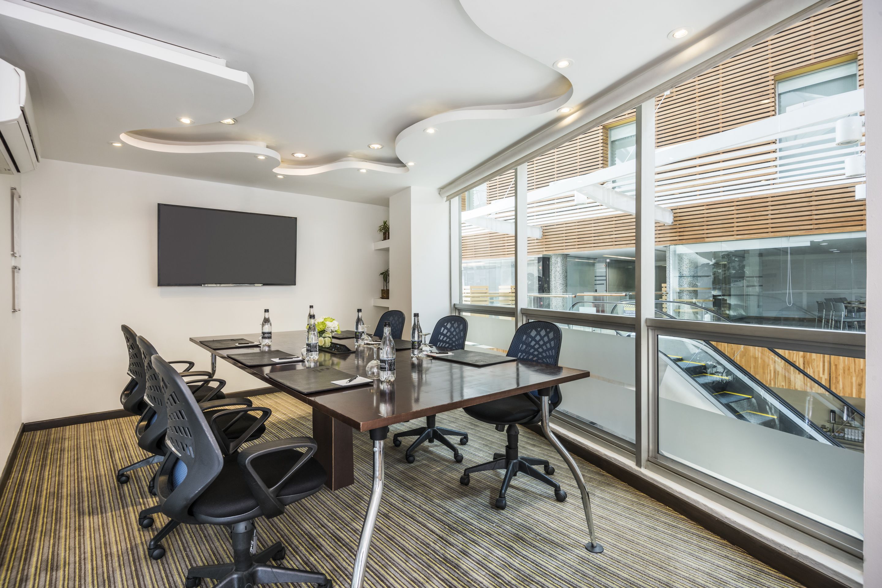 A modern conference room with a large table, four chairs, a flat-screen TV, and large windows with a view of the building's interior.