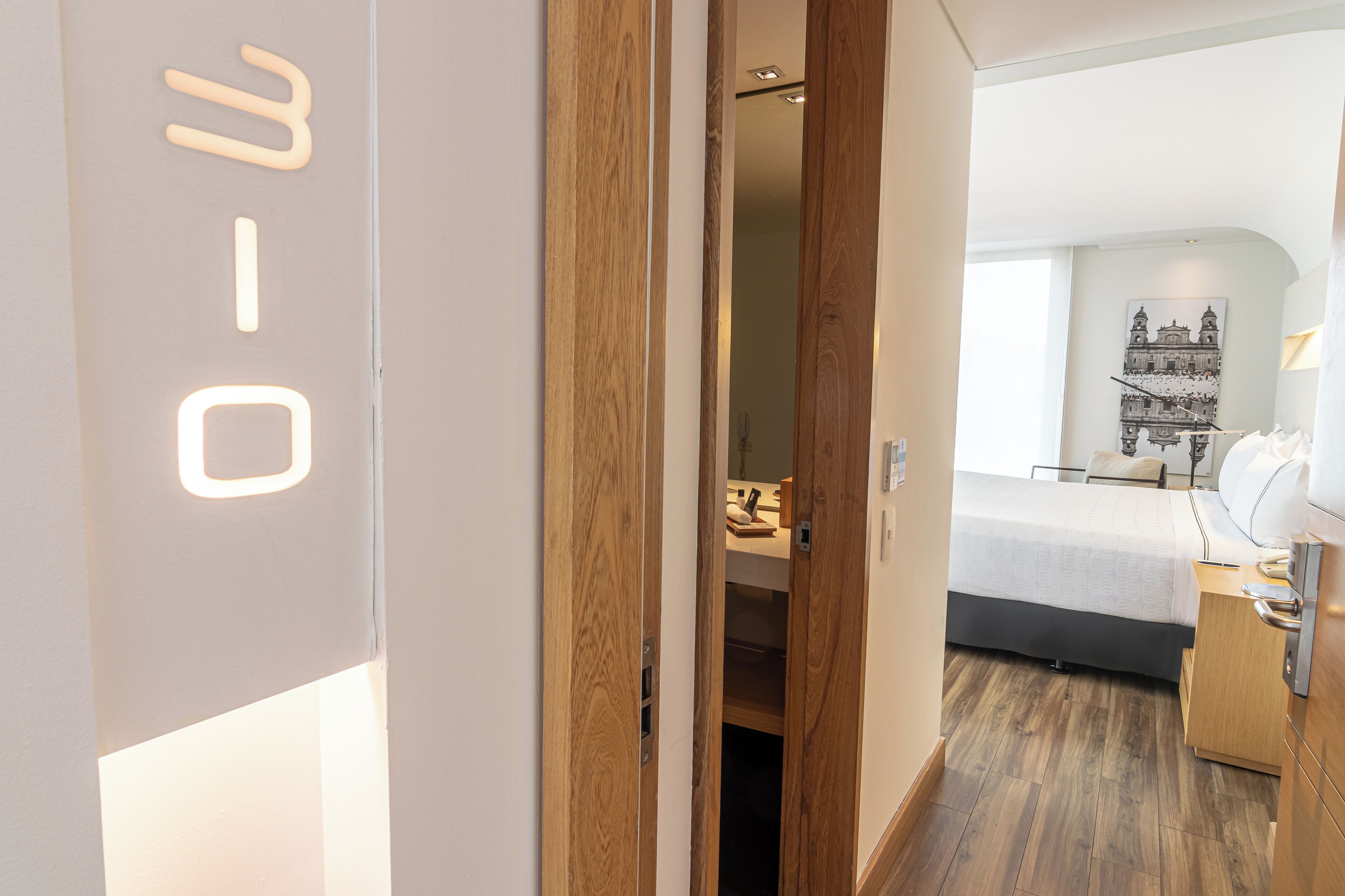 Een moderne hotelkamer met een houten deur, een bed met een wit hoofdeinde, een nachtkastje met een lamp en een aan de muur bevestigd telefoon.