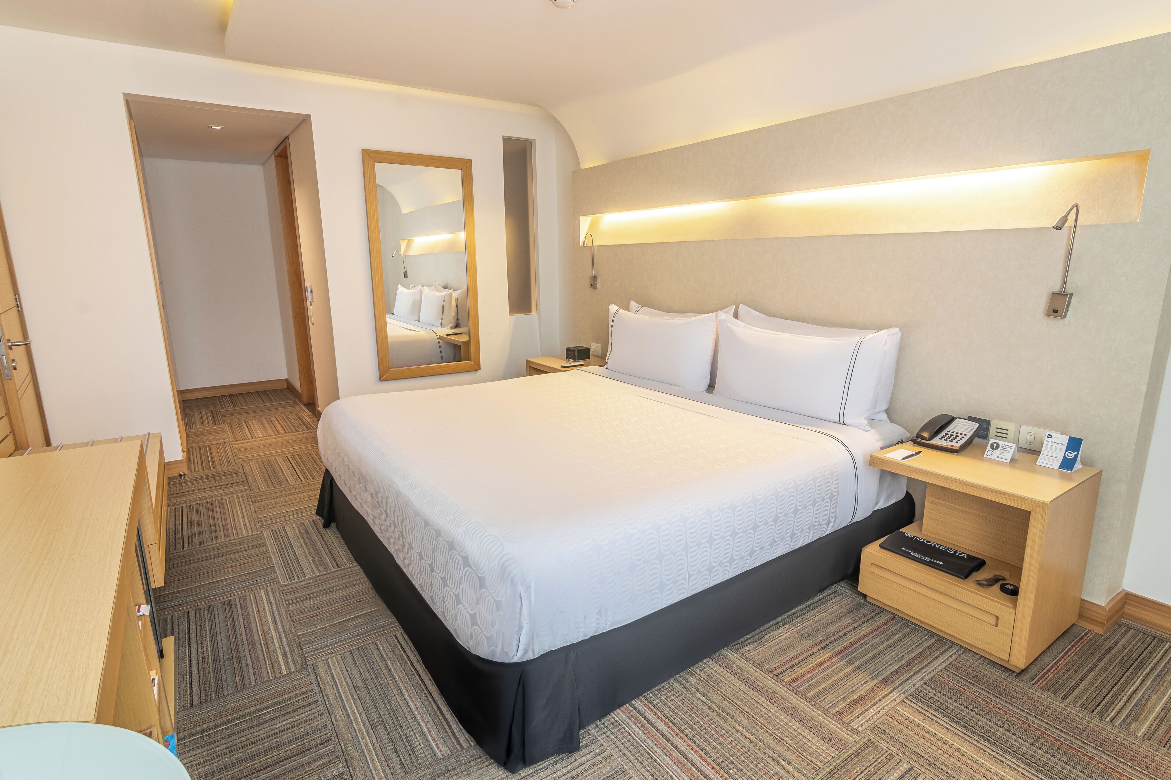 A hotel room with a large bed, bedside tables, a mirror, and a wall-mounted light fixture.