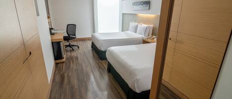 A hotel room with two beds, a desk with a chair, a computer, and a window providing a view.