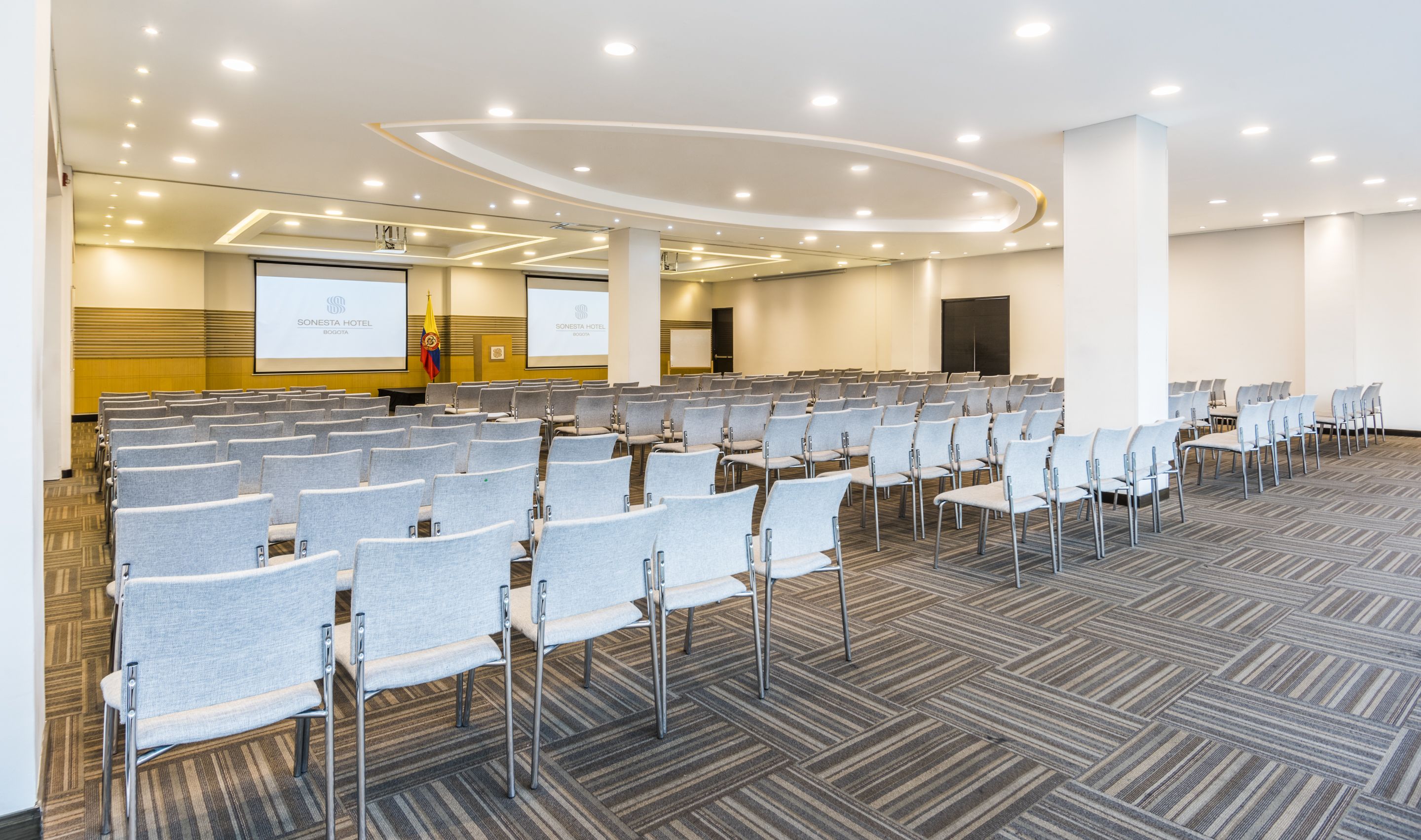 Sala de conferências com fileiras de cadeiras cinzas, piso carpeteado e tela para apresentação na frente.