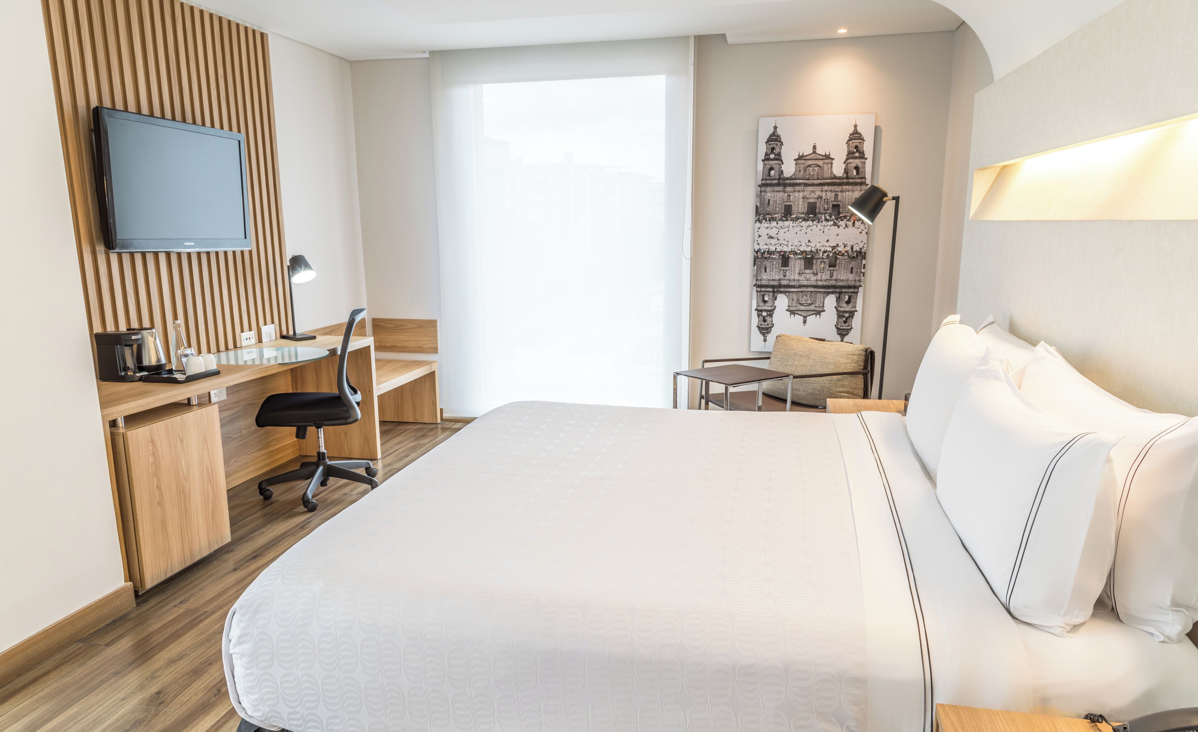 Una habitación de hotel moderna con una cama grande, un escritorio con una silla, un televisor de pantalla plana y un ventanal con vistas a un edificio.