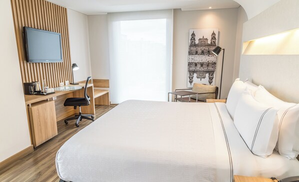 Una habitación de hotel moderna con una cama grande, un escritorio con una silla, un televisor de pantalla plana y un ventanal con vistas a un edificio.