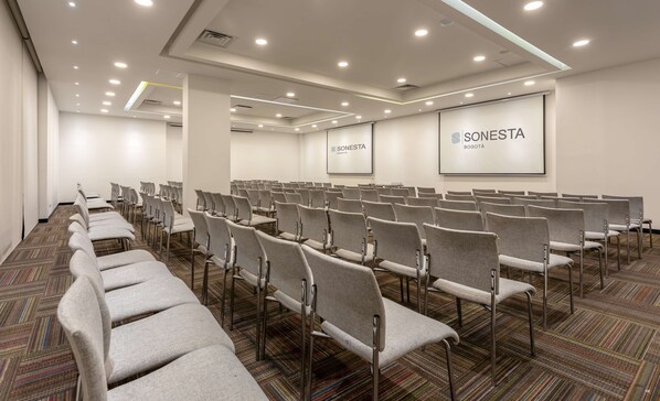 Un salón de conferencias con hileras de sillas grises, piso alfombrado e iluminación en el techo.