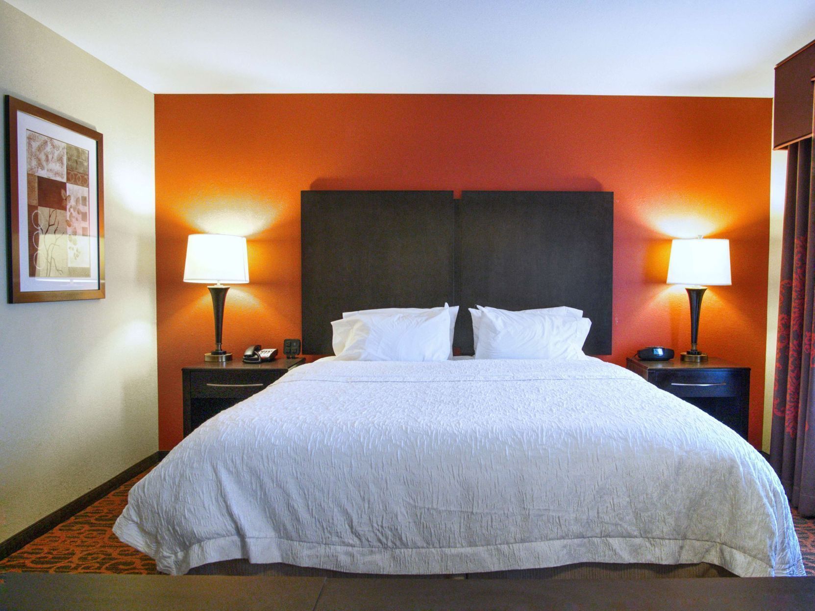 Una camera d'albergo con un letto grande, due lampade ai comodini, un quadro appeso alla parete e un tappeto rosso.