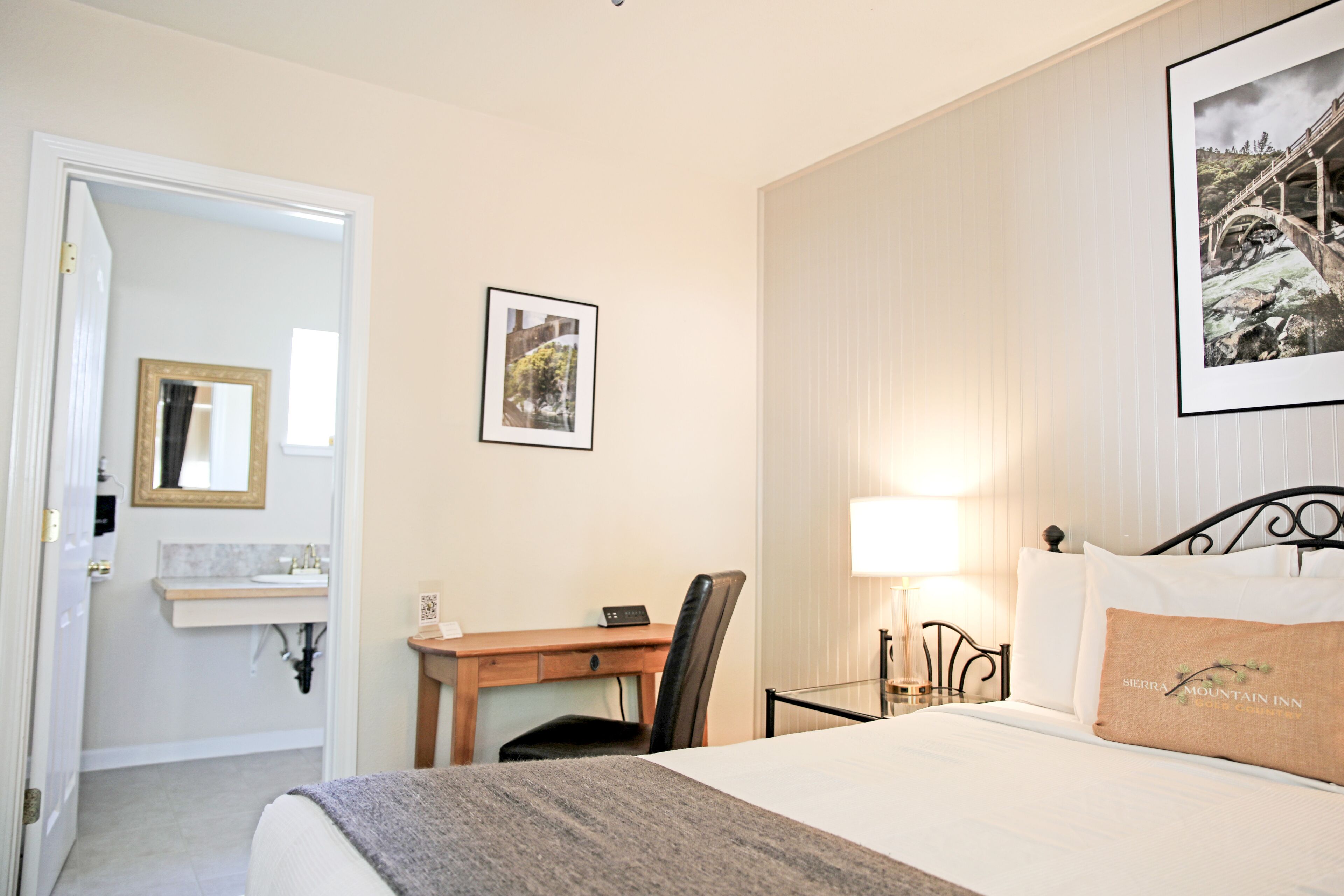 Habitación de hotel con cama, escritorio, silla, mesita de noche con lámpara, un cuadro en la pared y un baño visible a través de una puerta abierta.