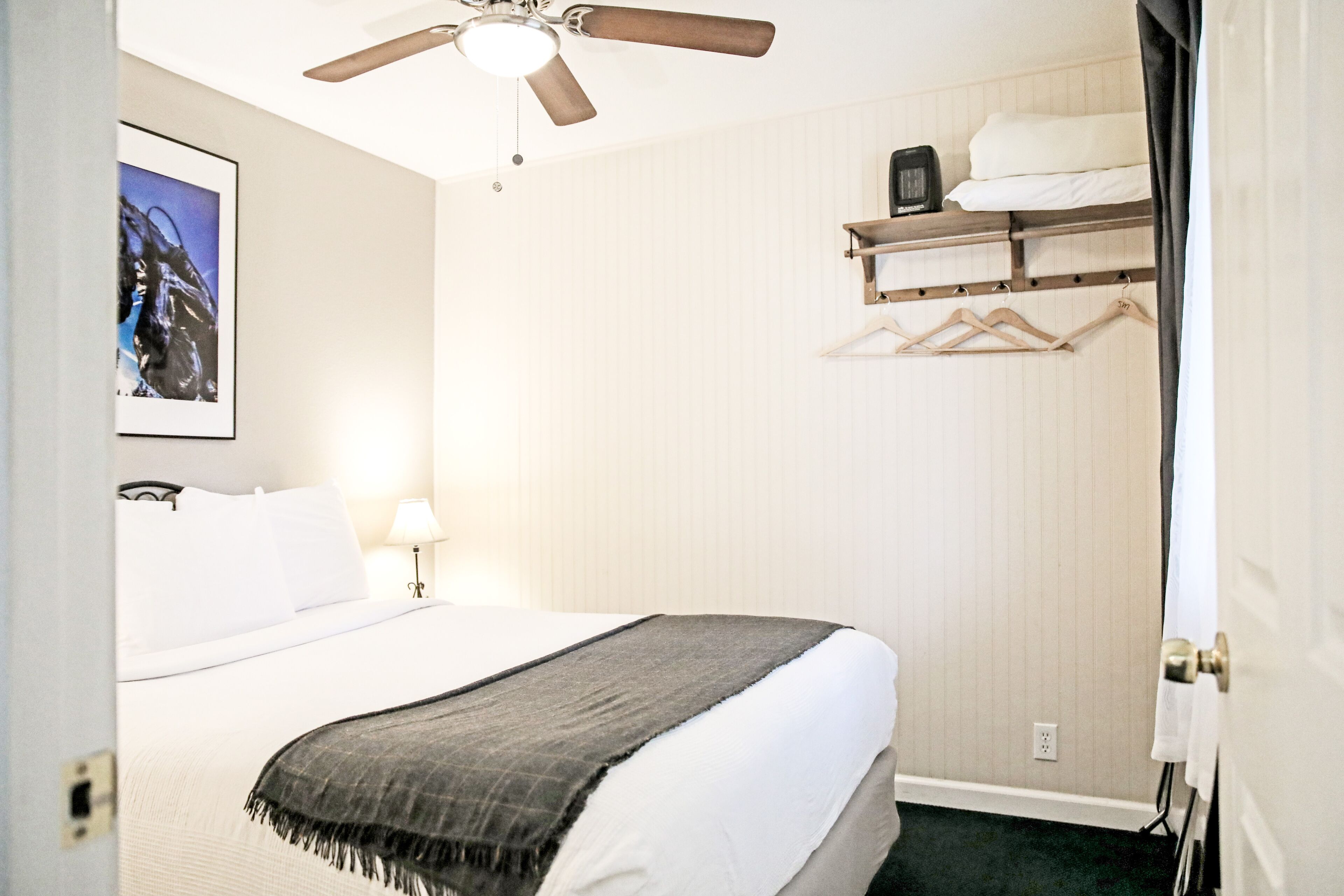 Una habitación de hotel con una cama, un ventilador de techo, una mesita de noche con lámpara, un televisor montado en la pared y un perchero.