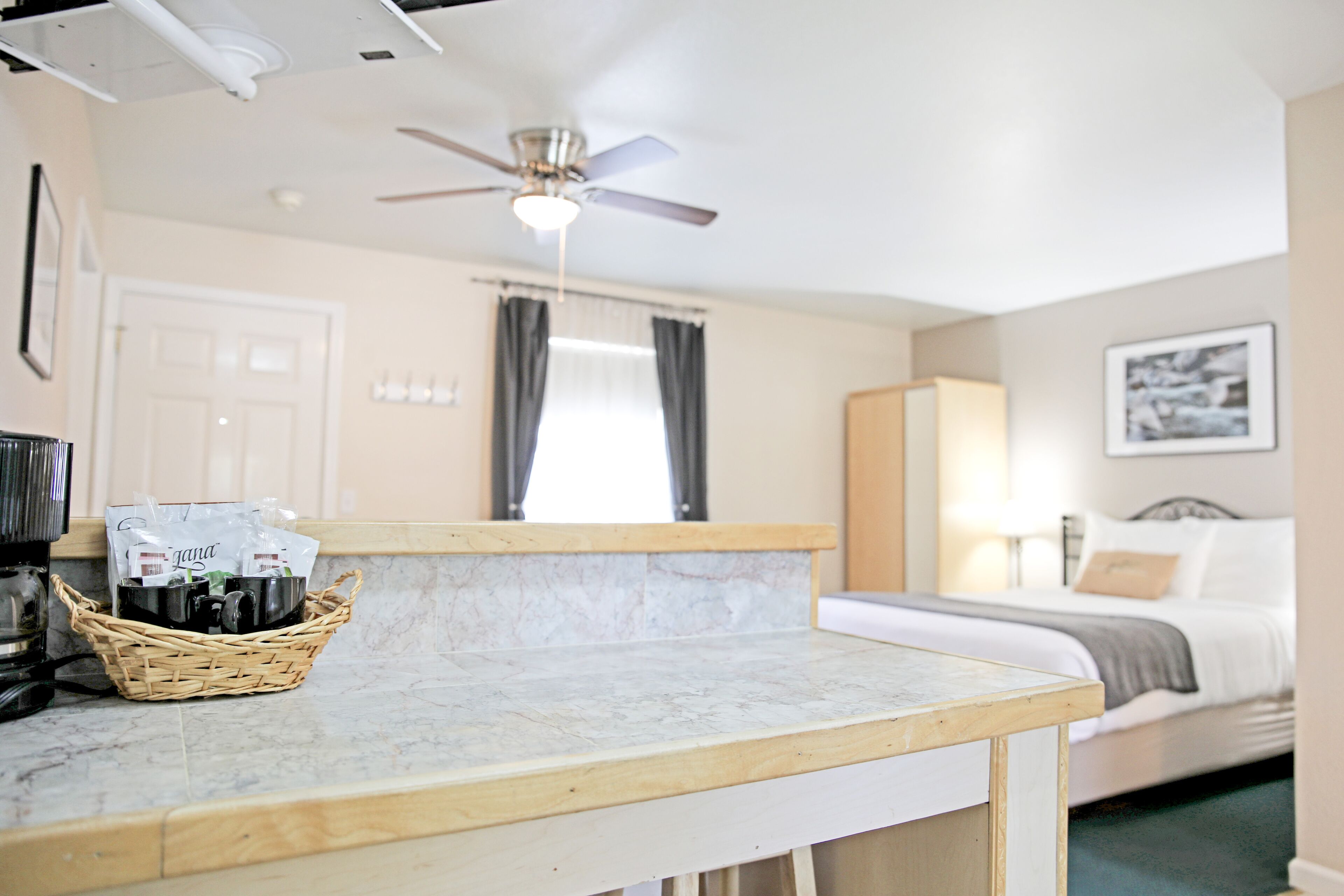 A hotel room with a bed, a bedside table with a basket, a ceiling fan, and a window with curtains.