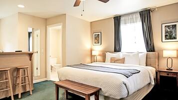 A hotel room with a bed, a desk with two stools, a bench, a ceiling fan, and a window with curtains.