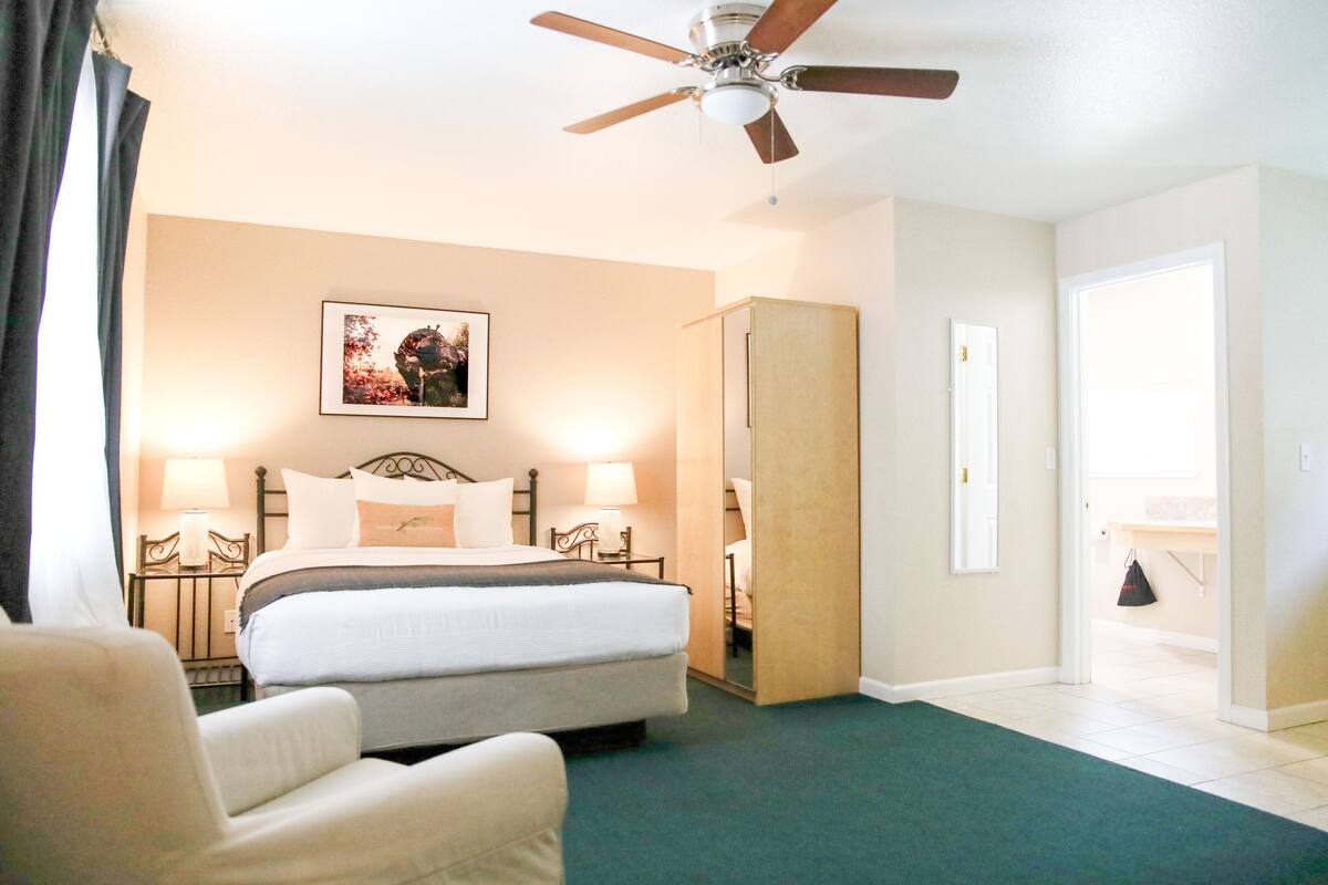 A hotel room with a bed, two bedside lamps, a ceiling fan, a large mirror, and a chair.