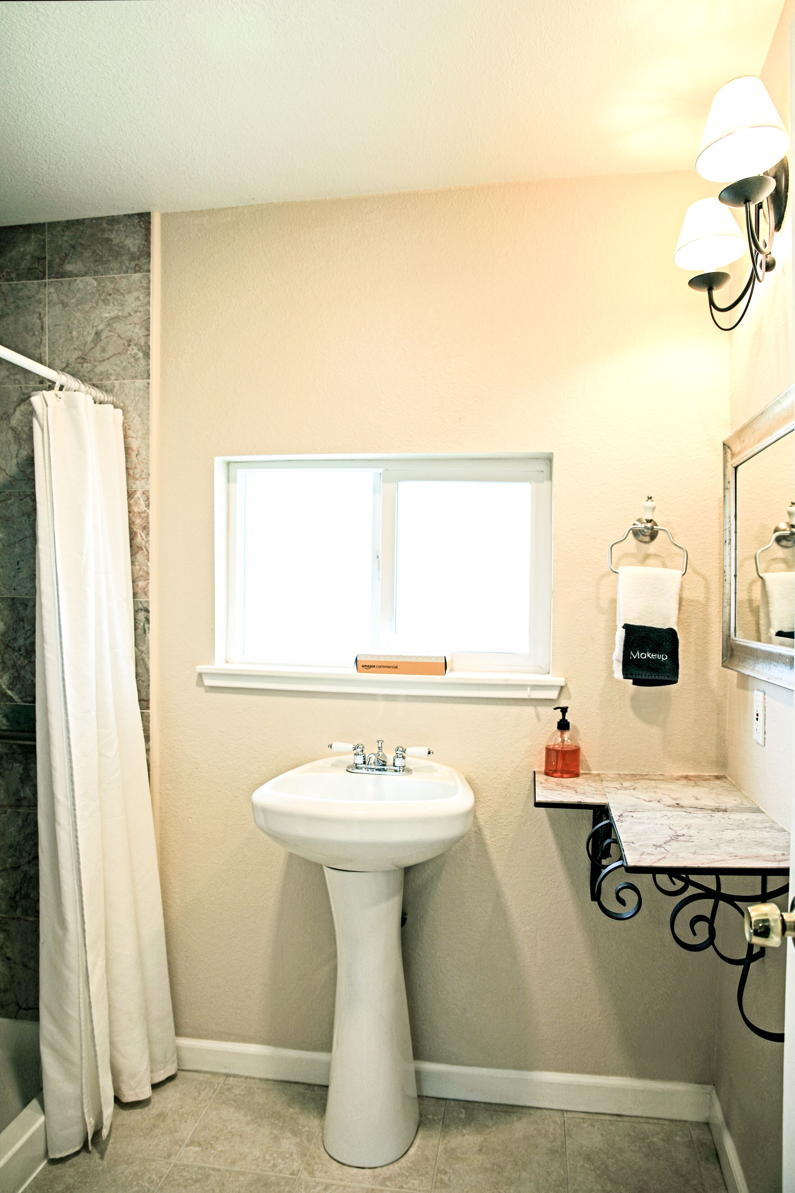 A bathroom with a pedestal sink, a window above the sink, a towel rack with a towel, a soap dispenser, and a mirror with lights.