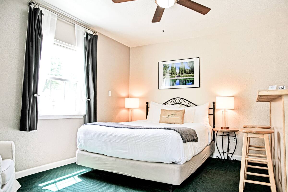 A neatly arranged bedroom with a bed, bedside lamps, a ceiling fan, a window with curtains, and a framed picture on the wall.