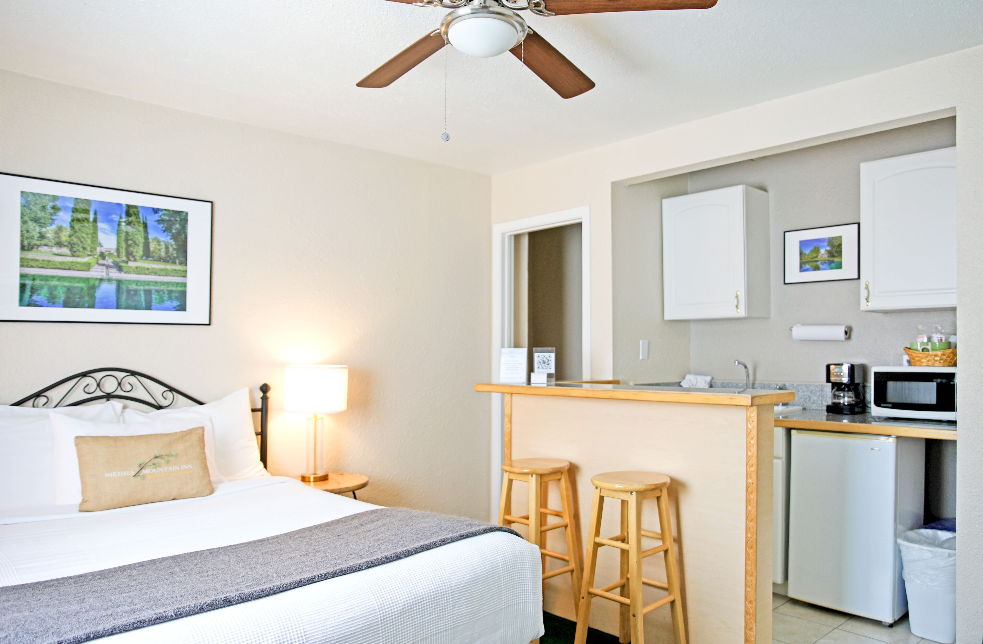 Habitación de hotel con cama, escritorio con taburetes, microondas, refrigerador y ventilador de techo.