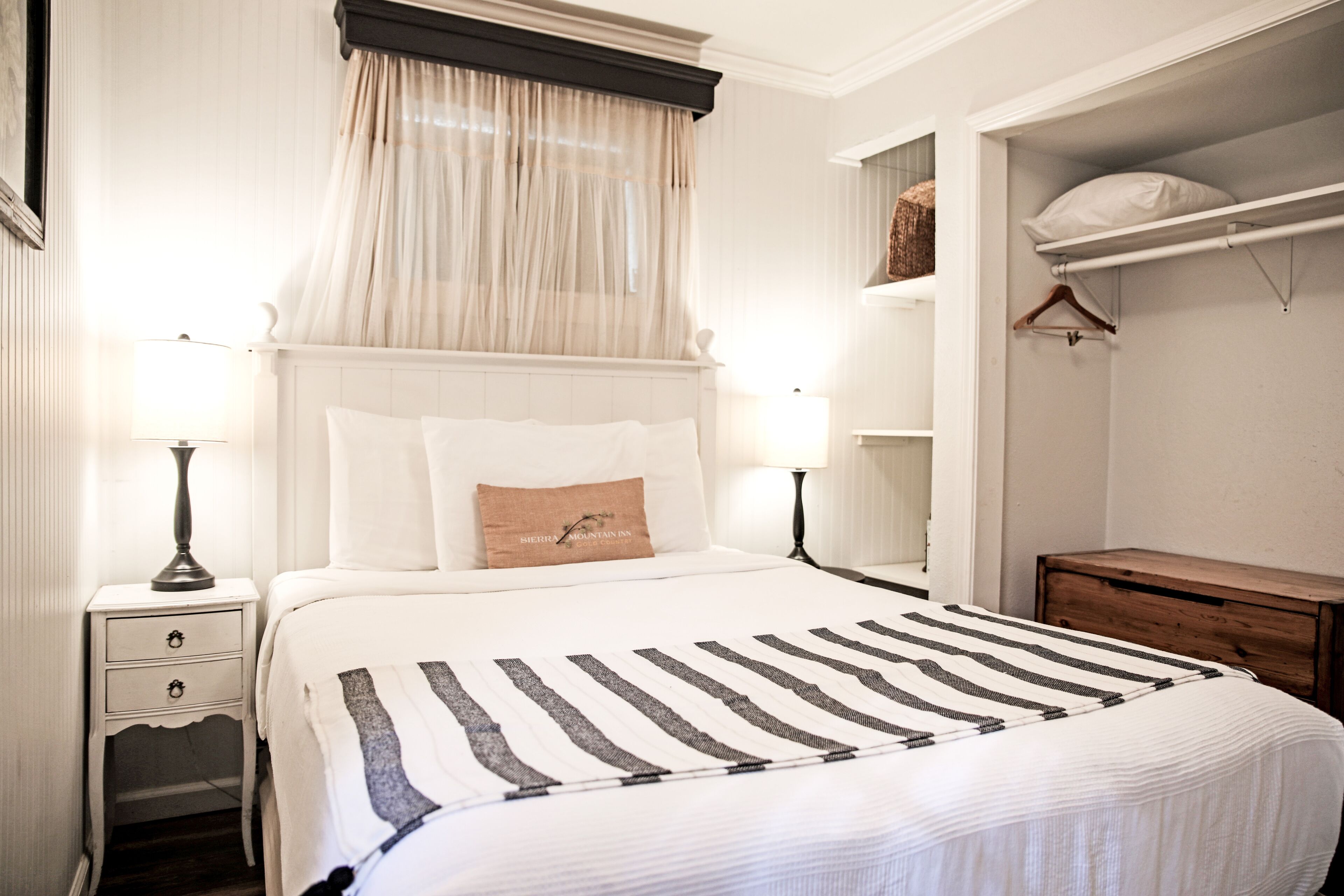 Una cama bien tendida con una colcha a rayas, una mesita de noche con lámpara, un tablero auxiliar con cajones, una ventana con cortinas traslúcidas y un aparador de madera con una cesta colgante.