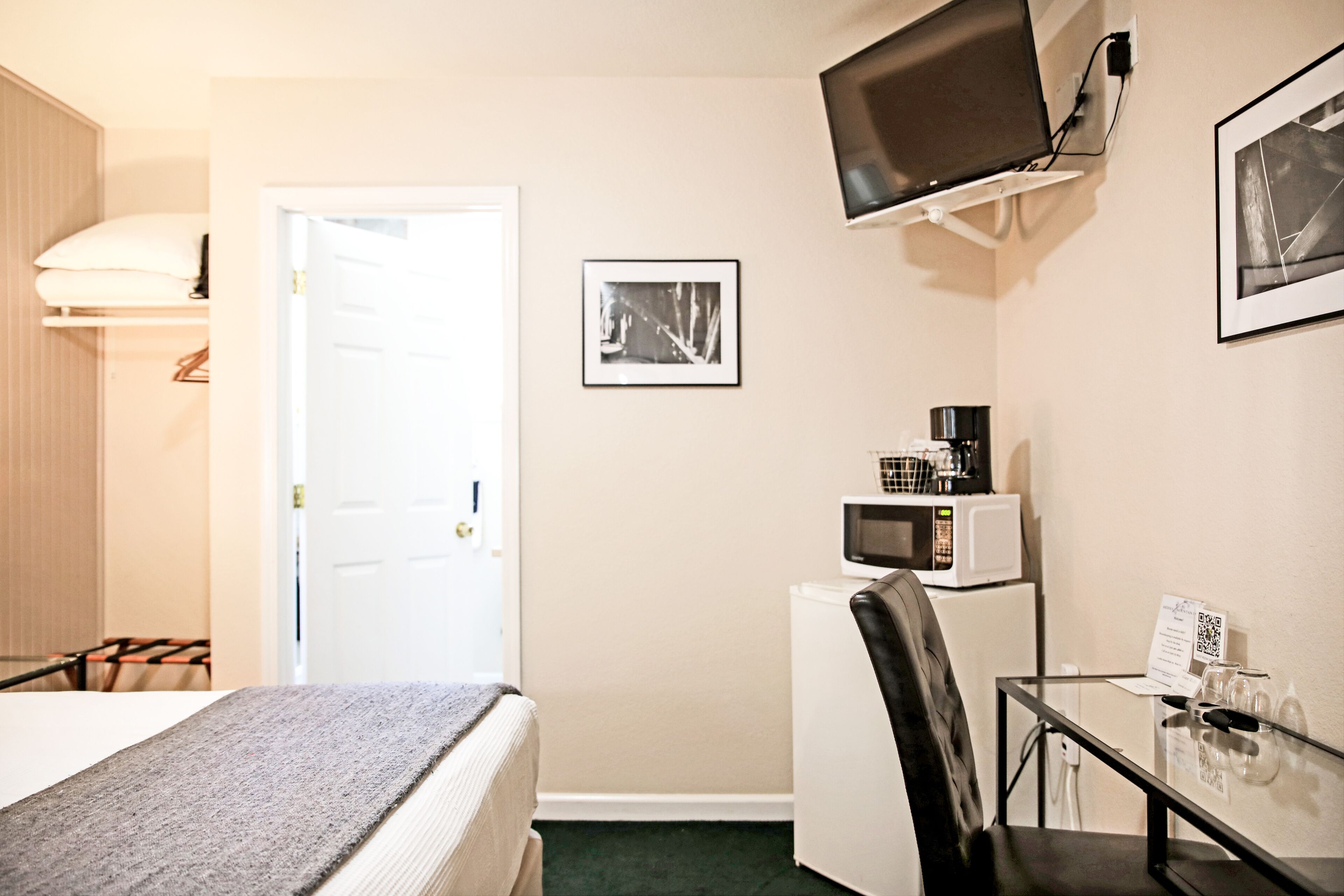 Ett kompakt hotellrum med en säng, ett skrivbord med en stol, en mikrovågsugn och en tv som är monterad på väggen, samt en dörr som leder till ett annat rum.