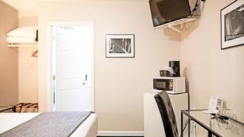 Ett kompakt hotellrum med en säng, ett skrivbord med en stol, en mikrovågsugn och en tv som är monterad på väggen, samt en dörr som leder till ett annat rum.