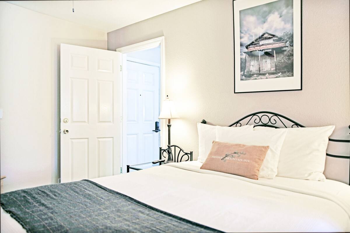 A hotel room with a bed, a nightstand, a lamp, a door, and a framed picture on the wall.