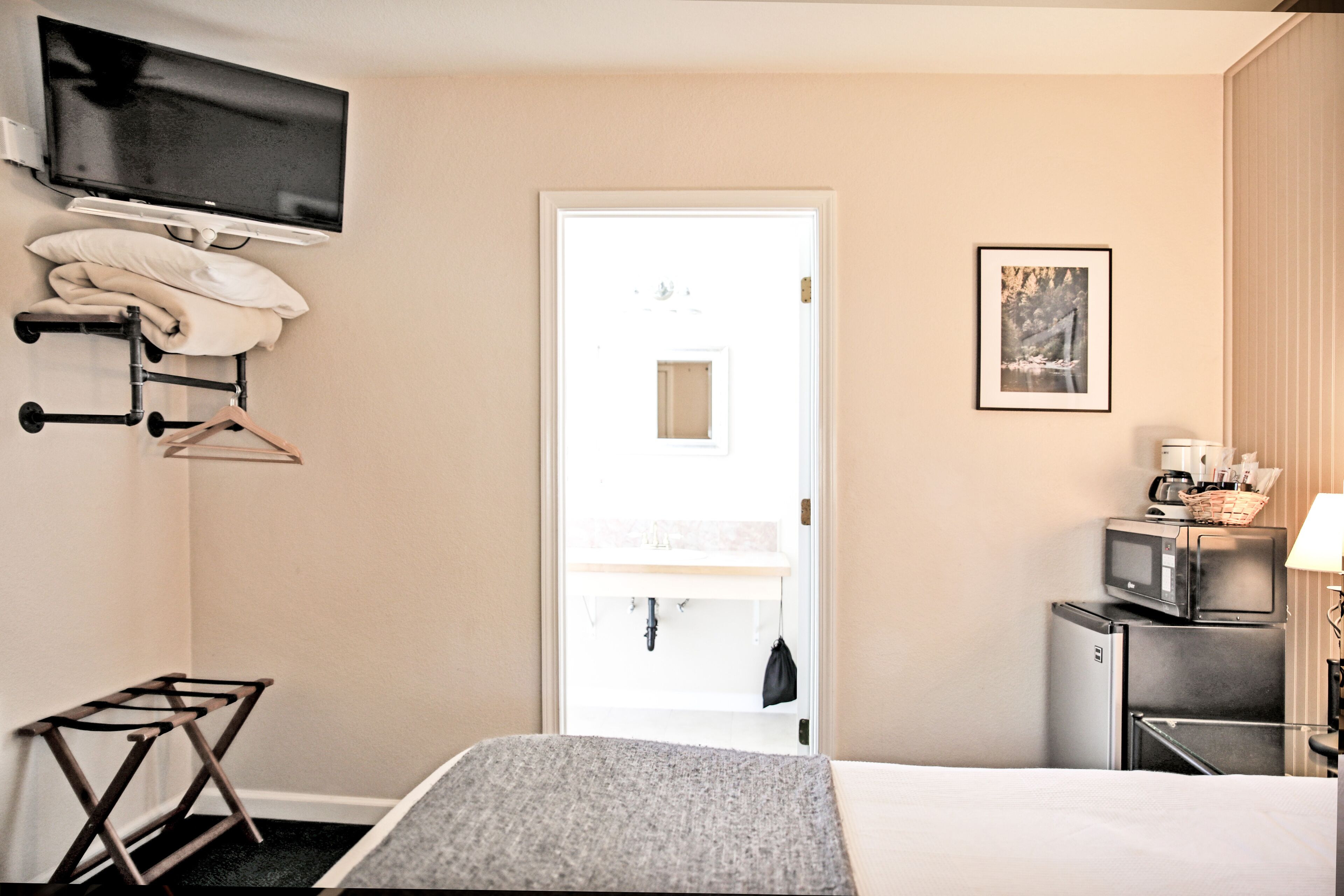 A hotel room with a bed, a TV mounted on the wall, a microwave, a refrigerator, a small table with a lamp, and a door leading to a bathroom.