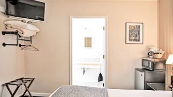A hotel room with a bed, a TV mounted on the wall, a microwave, a refrigerator, a small table with a lamp, and a door leading to a bathroom.