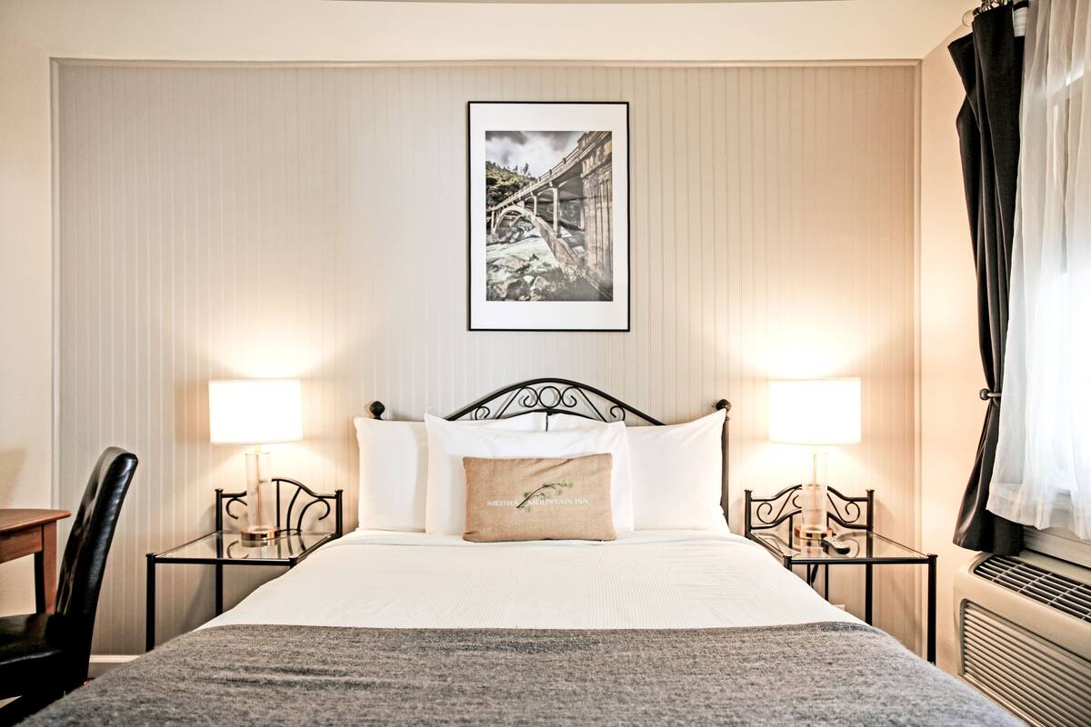 A hotel room with a bed, two bedside tables with lamps, a chair, a framed picture on the wall, and a window with curtains.